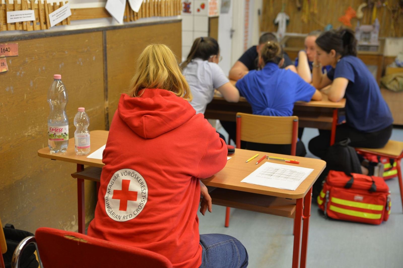 A student wearing a Red Cross hoodie sits at a desk among classmates in a classroom.