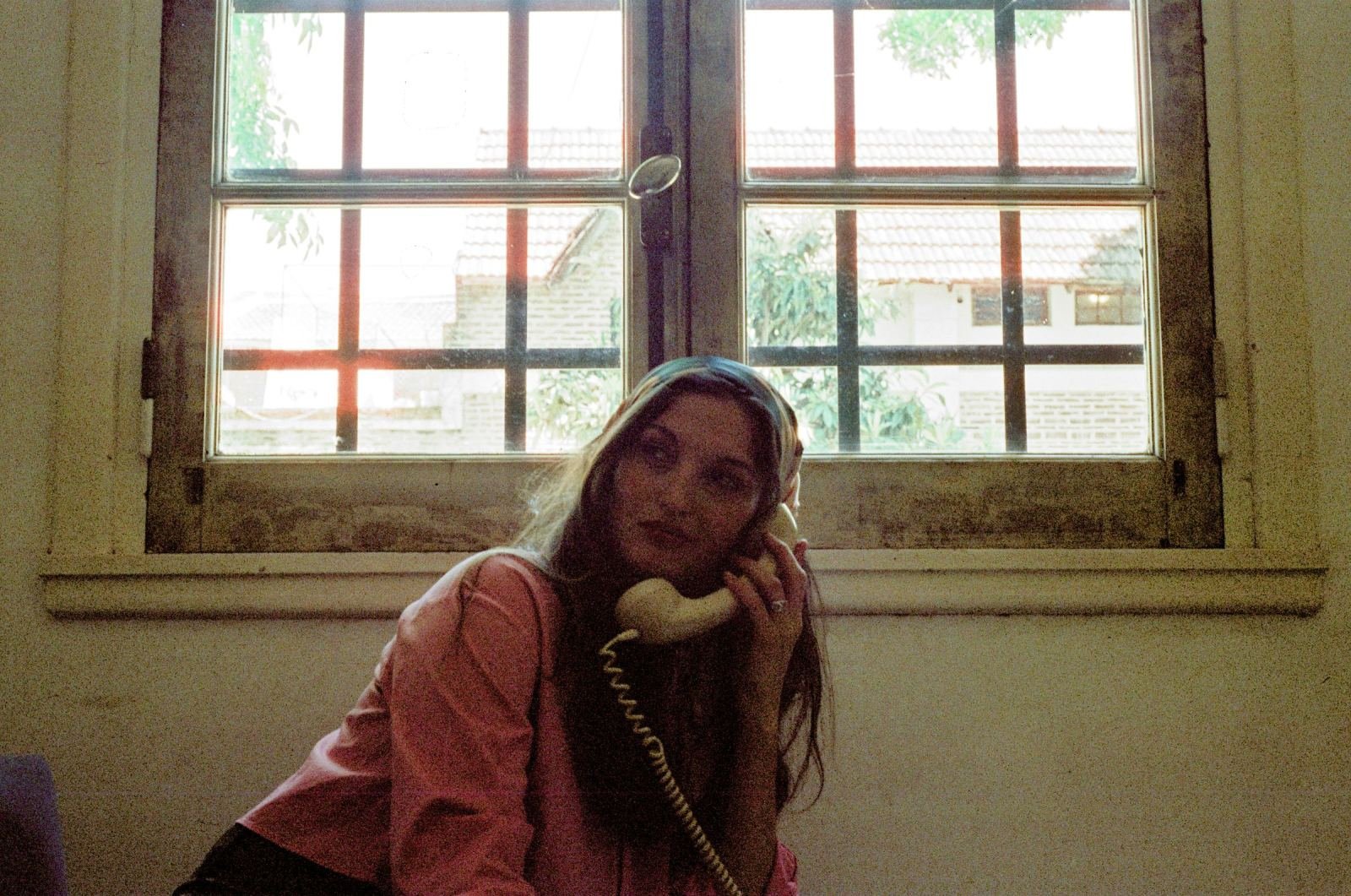 A woman in a pink shirt speaks into a vintage telephone receiver by a window