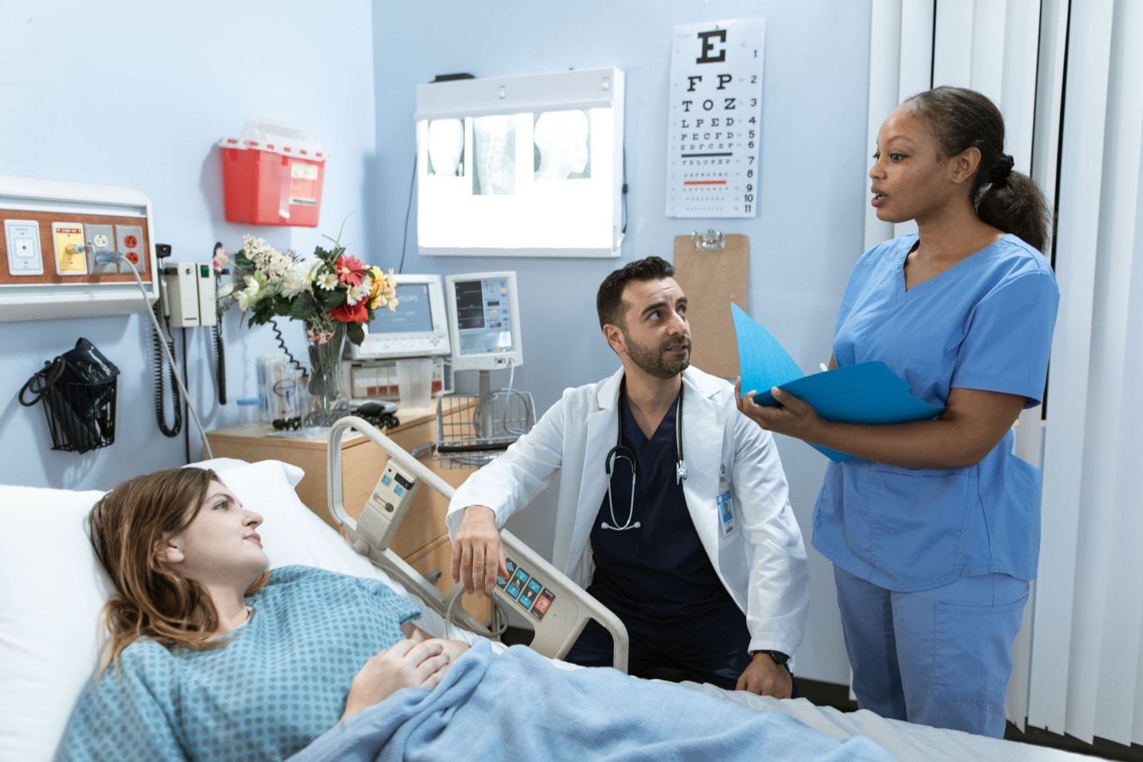 A doctor and nurse speak with a patient who is lying in a hospital bed