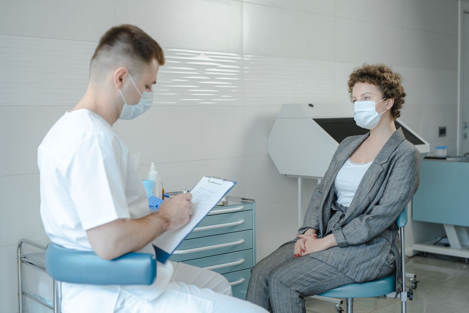 In a medical office, a masked clinician talks to a patient and writes on a clipboard.