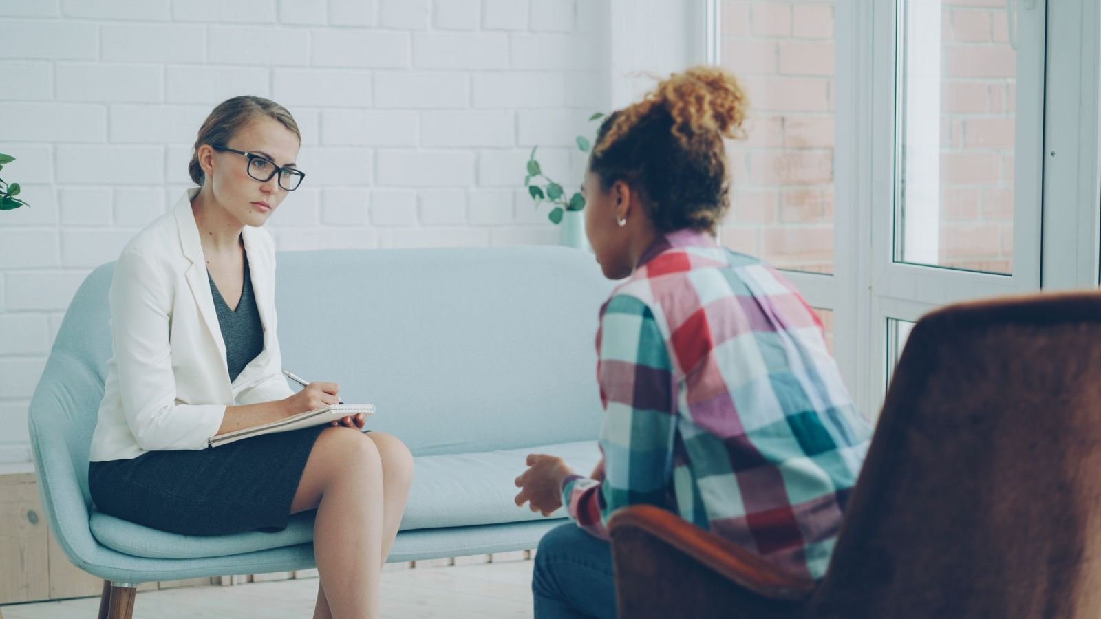 A client in a plaid shirt talks with a counselor in a white blazer, who writes notes.