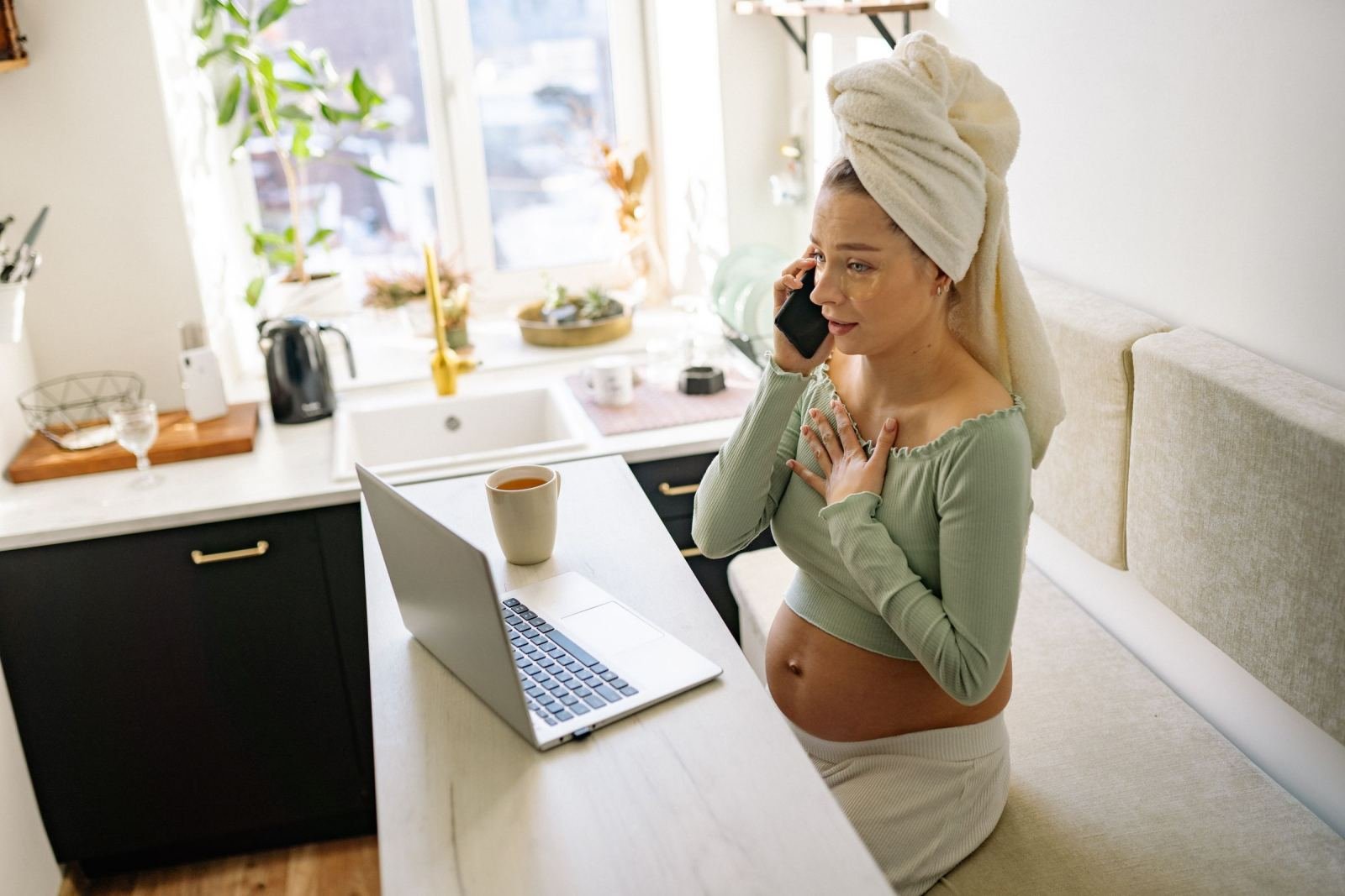 A pregnant woman holds a phone in her kitchen. She has a towel on her head.