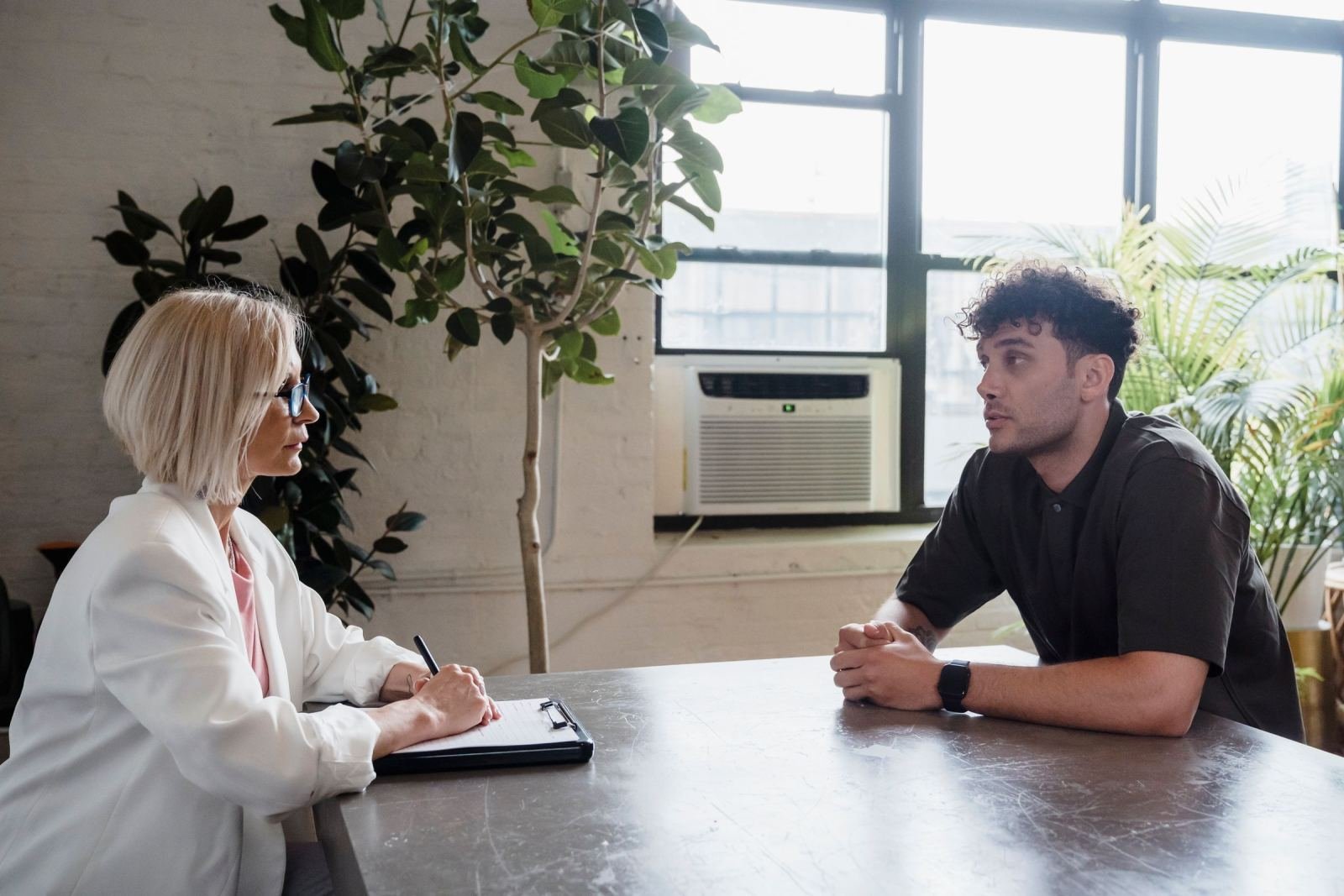 In a bright office, a man in a dark shirt sits across a table from a woman holding a clipboard. They are talking.