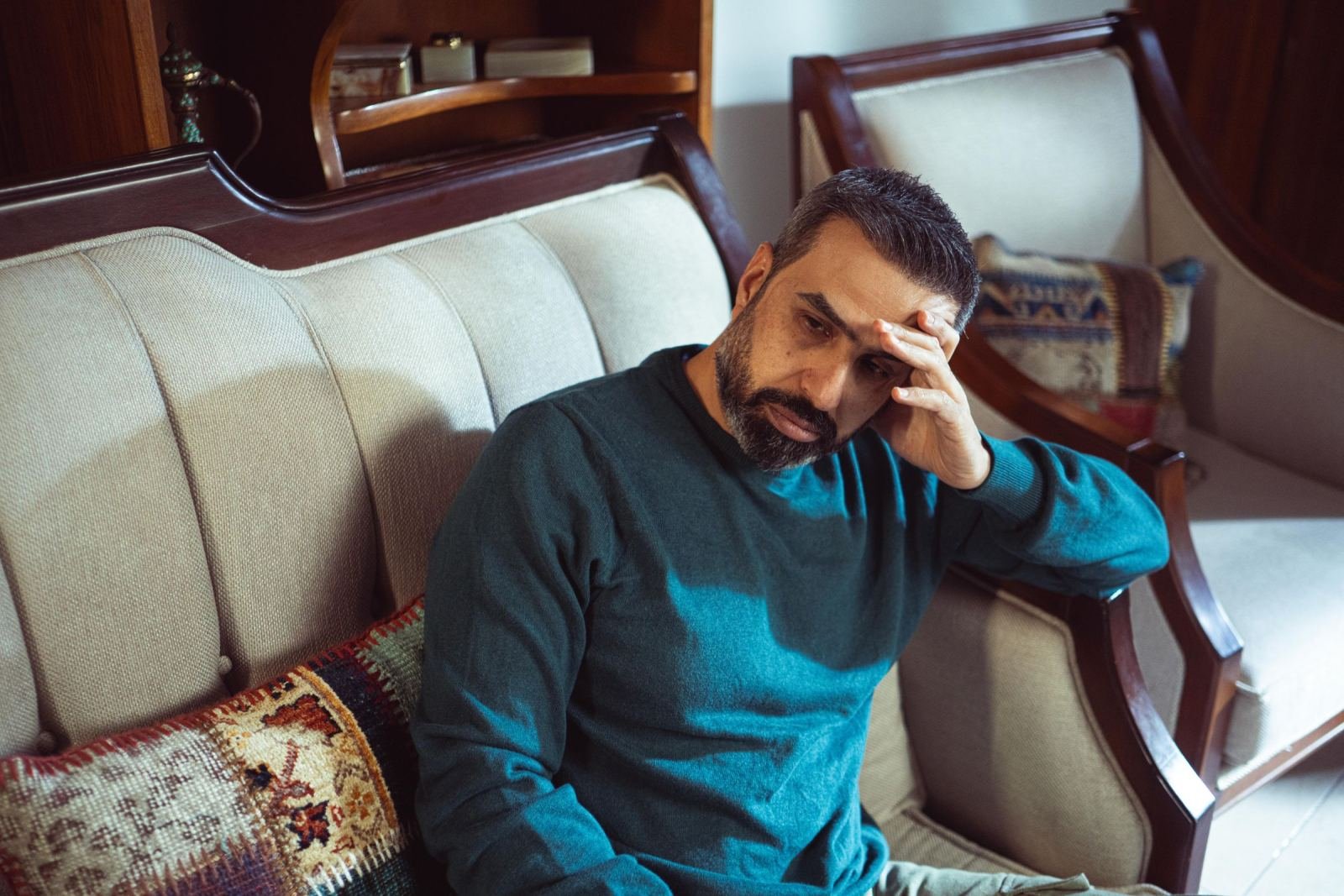 A man in a teal sweater sits on a couch, holding his head.