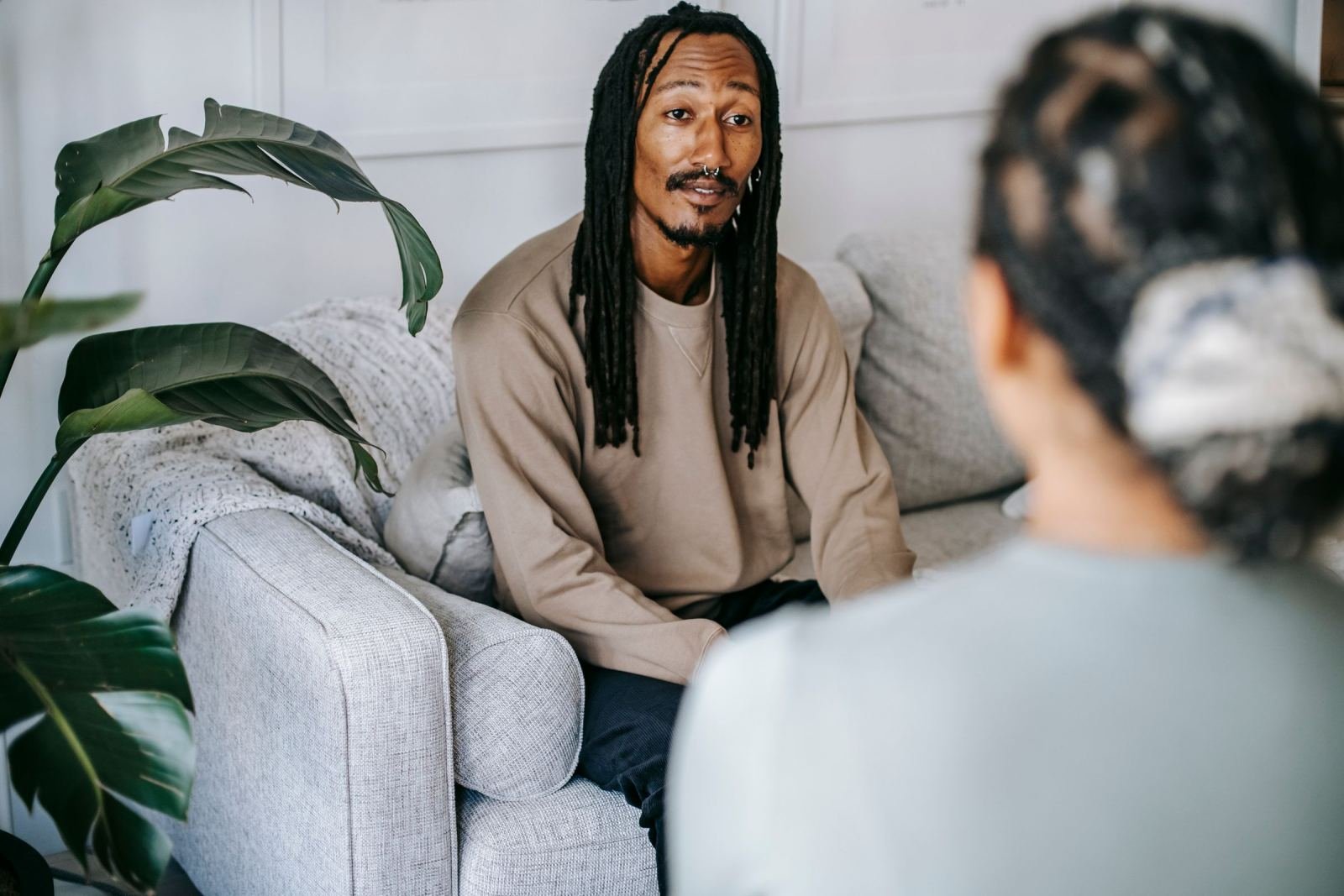 A person with dreadlocks is seated on a gray couch, speaking with someone else.