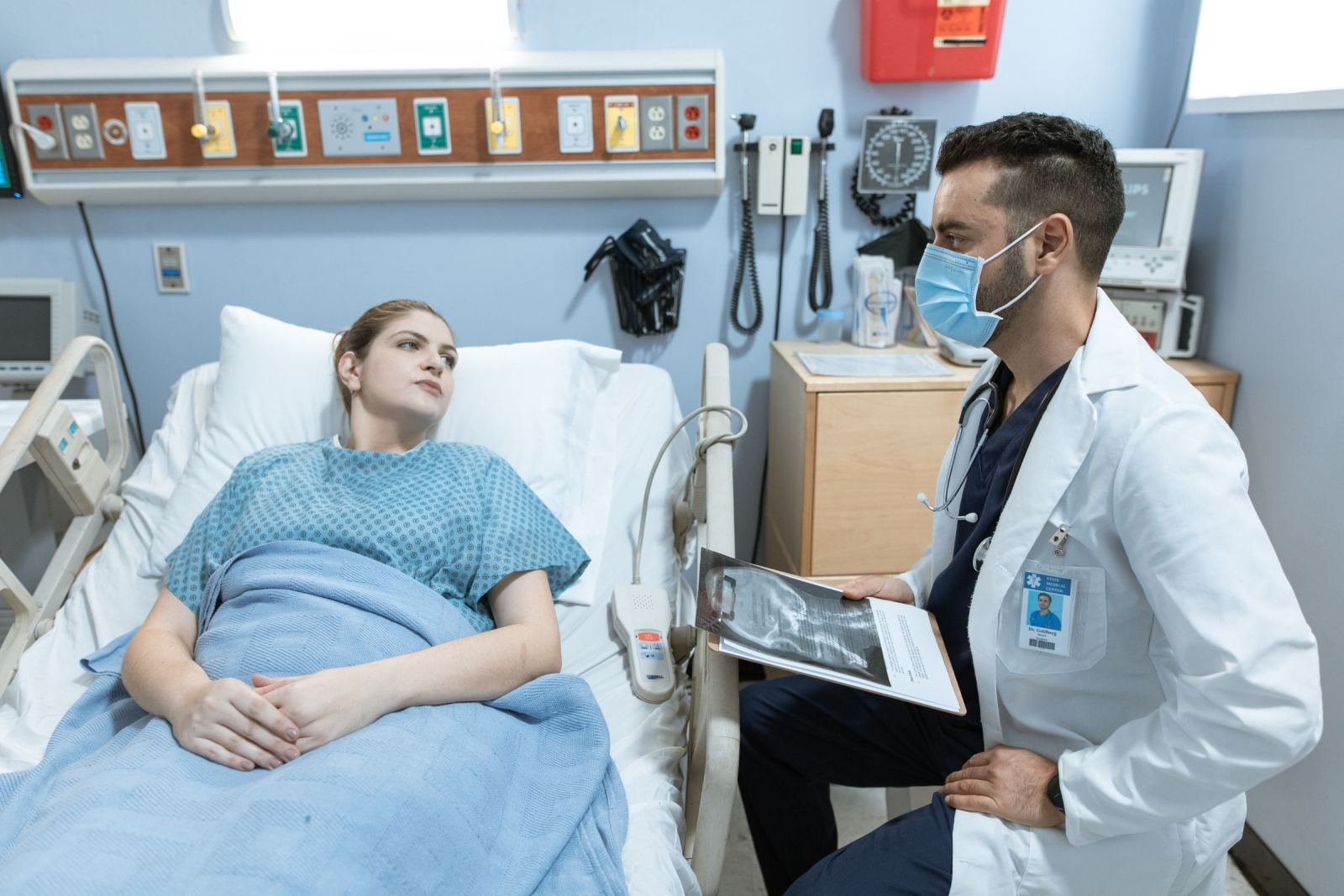 A doctor in a mask and white coat talks to a patient in a hospital bed