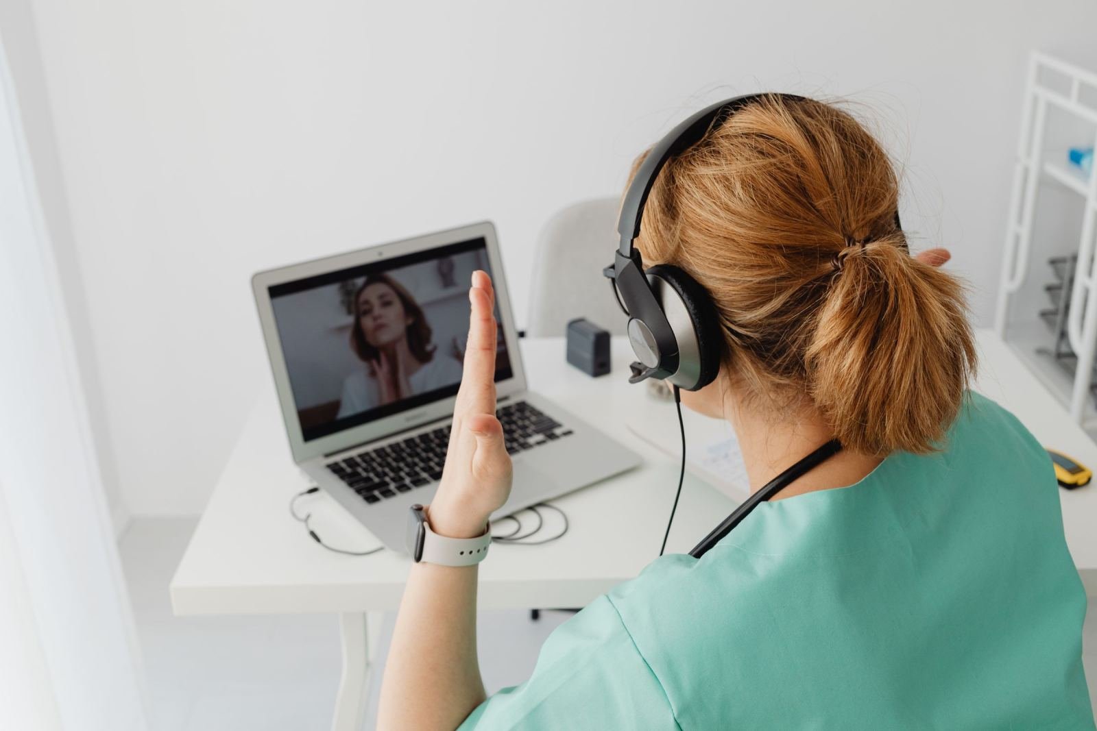 A woman in scrubs wears headphones and waves at someone on a laptop video call.