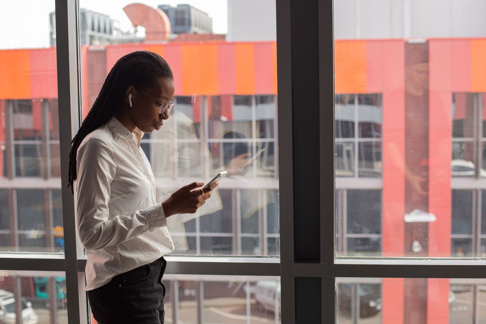 A woman in a white shirt stands by a window, looking at her phone.