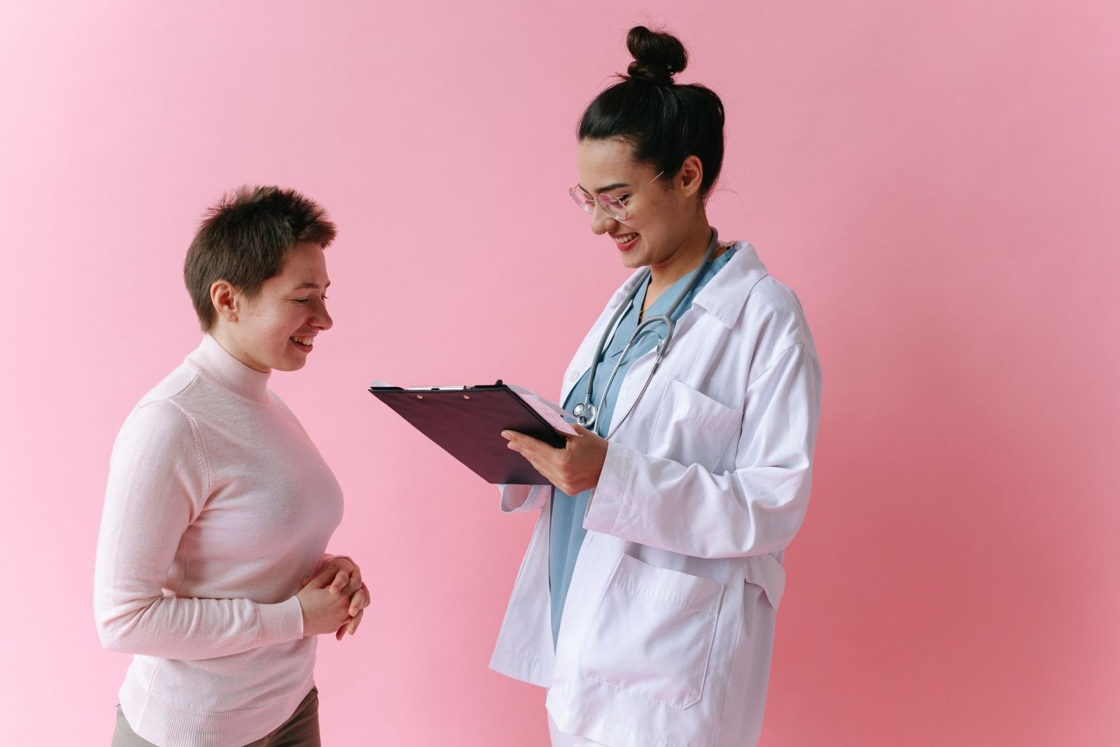 A healthcare provider in a white coat talks with a patient against a pink background