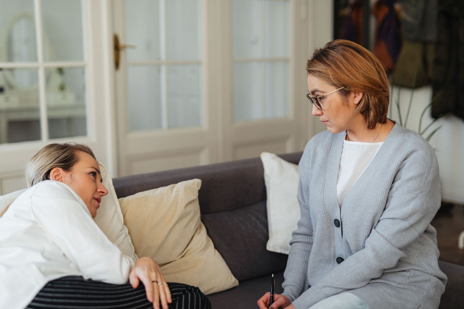 Two women talk in a bright office, one leaning on a sofa and the other holding a pen