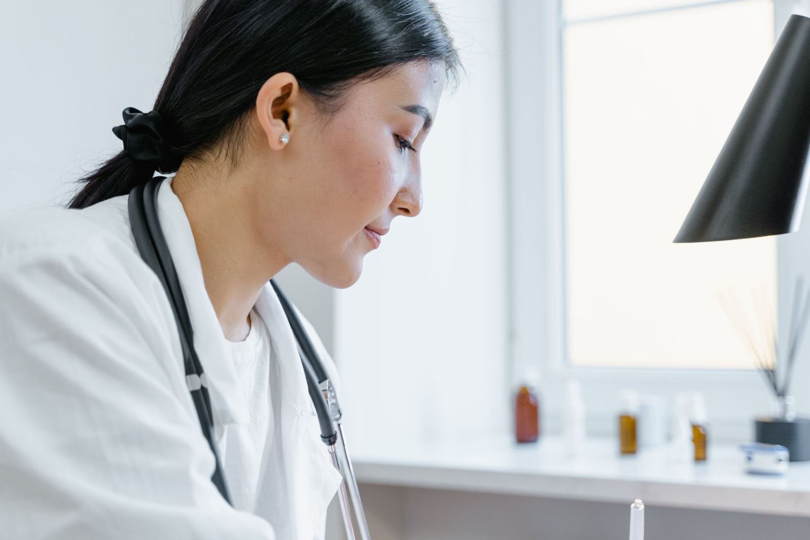 A doctor wearing a white coat and stethoscope looks down in a clinic.