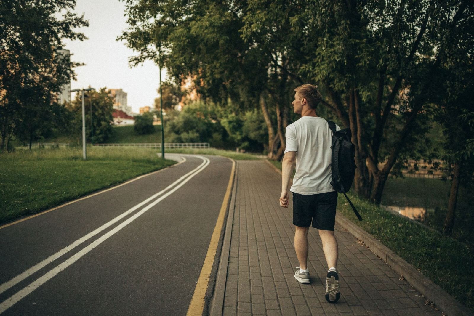 A man carrying a backpack strolls down a paved walkway in a green park.