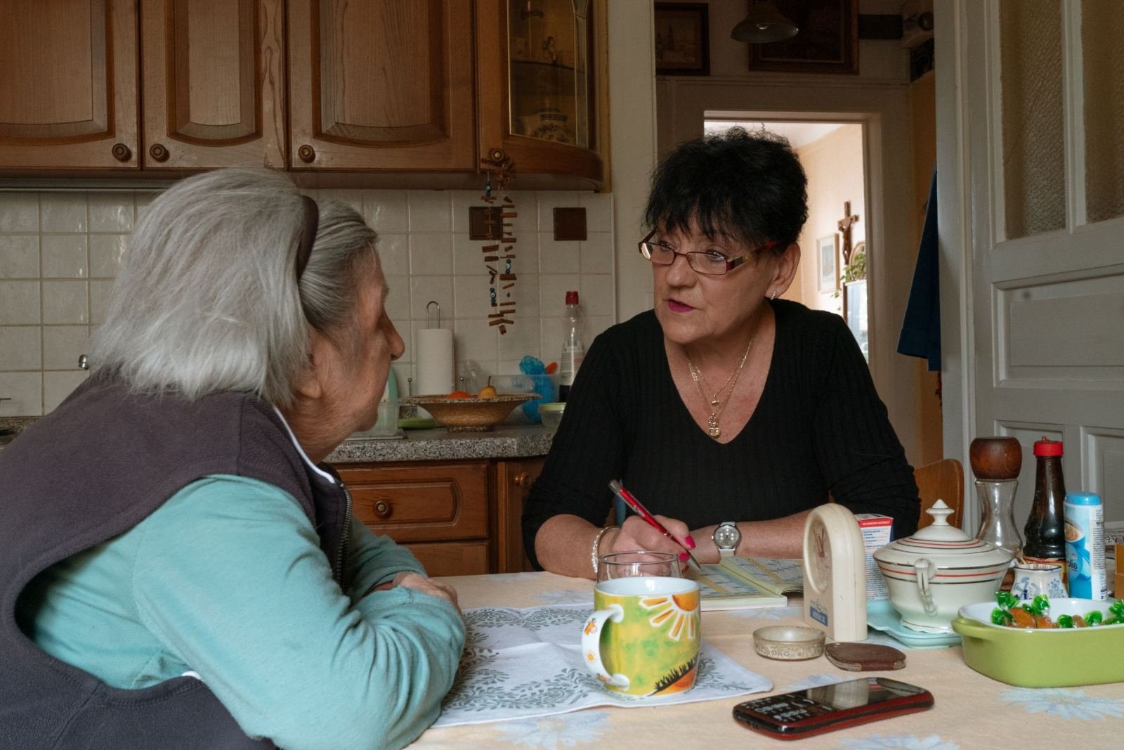Two women sit at a kitchen table talking while one holds a pen and notebook