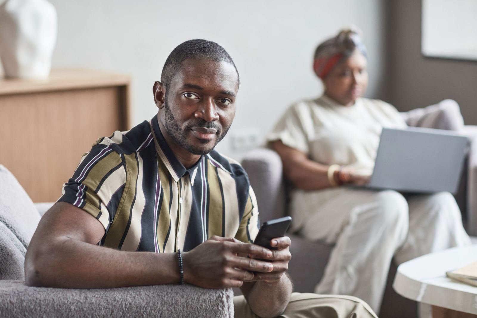 A man sits on a sofa in a bright living room, holding a smartphone