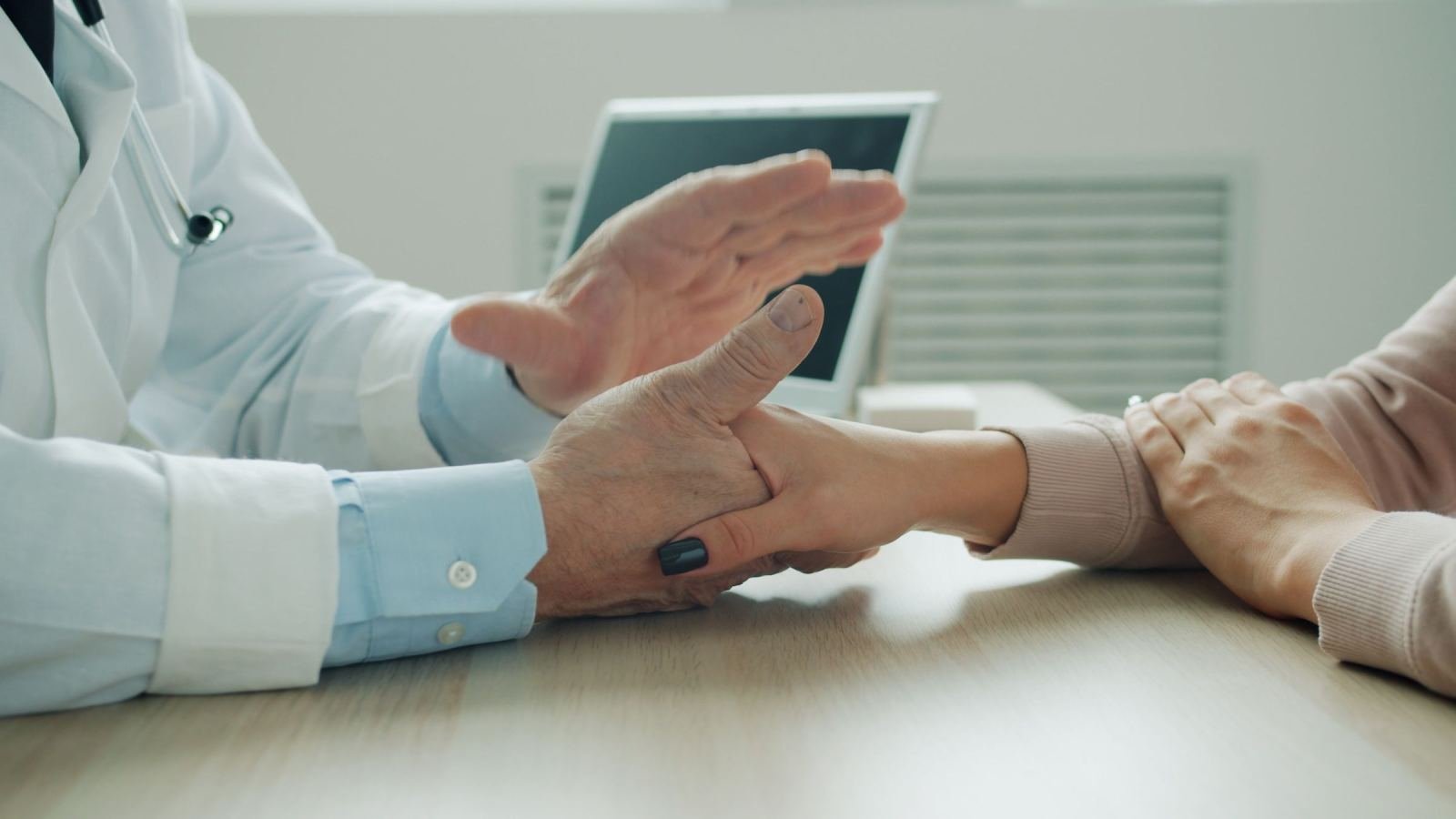 A clinician wearing a white coat holds a patient's hand across a desk