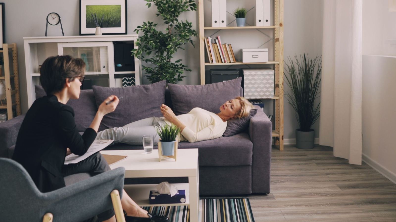 A woman sits back on a gray couch while a counselor in a dark jacket speaks with her.