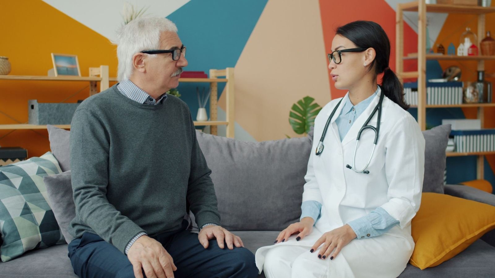 An older man sits on a sofa talking with a doctor in a white coat.