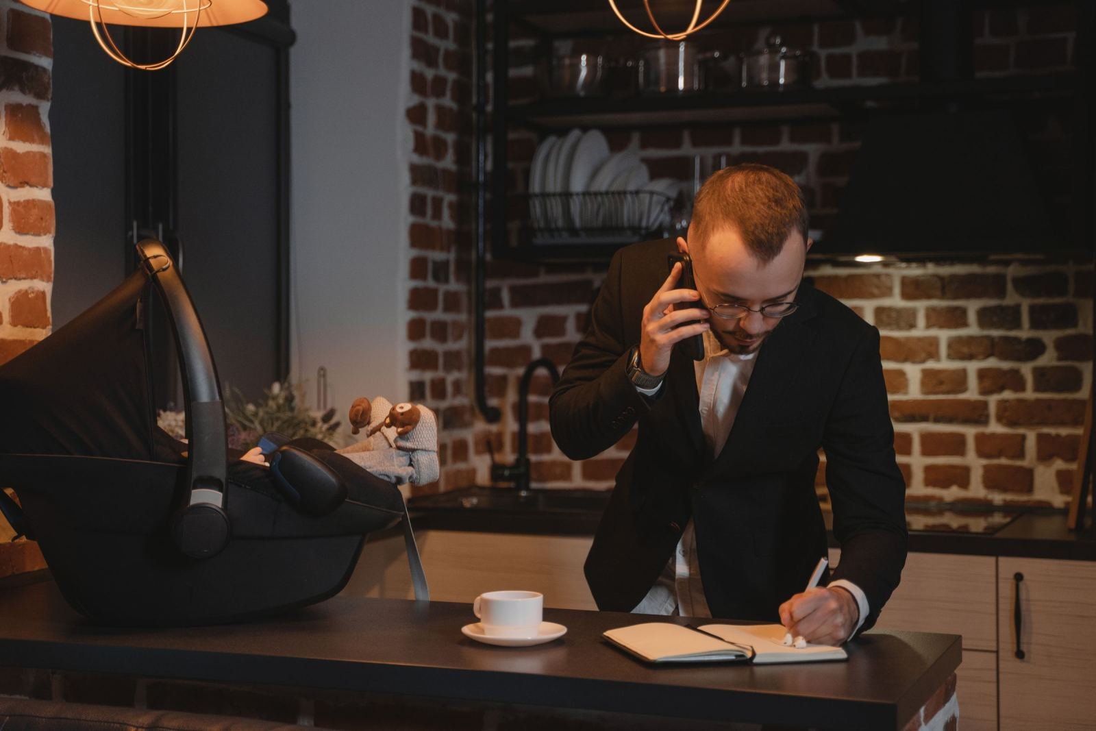 A man in a suit talks on the phone and writes in a notebook next to a baby car seat
