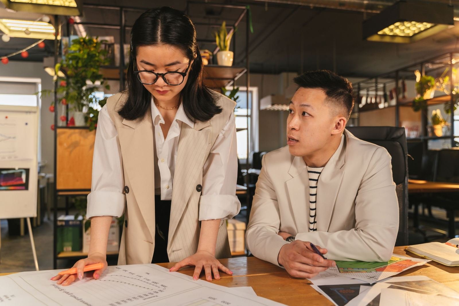 At a shared office desk, two coworkers look over papers and a calendar.